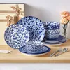 A set of blue and white patterned porcelain tableware is displayed on a wooden surface, with gift boxes and flowers in the background. Produktbild-Vorschau