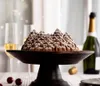 A Bundt cake shaped like a pine forest, dusted with powdered sugar, is presented on a dark wooden cake stand. Two glasses of champagne and a bottle of champagne are visible in the background, along with festive string lights. Anteprima dell’immagine prodotto