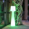 A tall, cone-shaped lamp with a white pleated shade is illuminated with green light, standing on a wooden deck next to a large, gnarled tree trunk. Produktbild-Vorschau