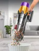 A hand holding a cordless vacuum cleaner, emptying debris into a trash can in a living room. Produktbild-Vorschau