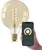 A hand holds a smartphone displaying a smart bulb control app next to a large, gold filament smart LED bulb. Produktbild-Vorschau