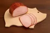 A piece of ham and three slices of ham are on a pig-shaped wooden cutting board, which is on a wooden table. Podgląd zdjęcia produktu