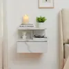 A white wall-mounted nightstand with a marble-like pattern is shown, featuring a drawer and two shelves. Decorative items like a candle, a bust, a plant, and books are placed on the shelves. Náhľad obrázku produktu