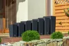 A row of black planters of varying heights are arranged on a patio next to a building and a wooden fence. Green bushes are in front of the planters. Anteprima dell’immagine prodotto