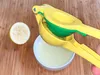 A yellow and green manual lemon squeezer is being used to squeeze a lemon into a white bowl. A halved lemon is visible next to the bowl. Produktbild-Vorschau