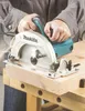 A person is using a Makita circular saw to cut a piece of wood on a workbench. Produktbild-Vorschau