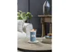 A lit candle in a blue glass jar sits on a round wooden table next to a stack of books and a small dish. A sofa and a plant are visible in the background. Anteprima dell’immagine prodotto