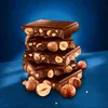 A stack of chocolate bars with hazelnuts, with loose hazelnuts scattered around the base. Náhľad obrázku produktu