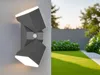 A modern outdoor wall light with a motion sensor is shown mounted on a wall, with a blurred background of a manicured garden and a fence. Anteprima dell’immagine prodotto