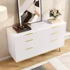 A white dresser with gold handles and legs, decorated with a framed abstract painting and a small bouquet of flowers. A record player is on top of the dresser. Podgląd zdjęcia produktu