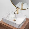 A close-up view of a rectangular marble-patterned vessel sink with a gold faucet, placed on a wooden countertop, with a round mirror reflecting the sink and faucet in the background. Náhľad obrázku produktu