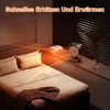 A compact heater is shown on a shelf next to books and a plant, with warm light emanating from it and illuminating a bed in a bedroom. Produktbild-Vorschau