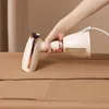 A hand holding a beige and rose gold garment steamer over a brown fabric surface. Produktbild-Vorschau