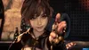 A close-up of a young woman with short brown hair, wearing a black outfit with cutouts, pointing her finger towards the camera with a playful expression. The background is blurred and appears to be an indoor or industrial setting. Produktbild-Vorschau