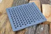 A square cast iron grate with many circular holes lies on a pile of chopped wood. Voir les images du produit