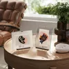 Two framed art prints are displayed on a wooden table, with a leather chair and greenery in the background. Produktbild-Vorschau