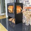 A modern black wood-burning stove with a fire burning inside, placed on a dark tiled hearth in a room with a brick wall and dried flowers in a vase. Produktbild-Vorschau