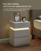 A white nightstand with LED lighting is shown next to a bed, with a framed photo and an alarm clock on top. Náhľad obrázku produktu