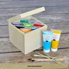 A wooden box filled with small paint tubes, with two larger paint tubes and brushes in front of it, on a wooden surface. Produktbild-Vorschau