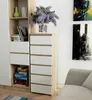 A modern dresser with white drawers and a wood-tone frame, placed next to a bookshelf with decorative items and books. Artwork hangs on the wall behind. Produktbild-Vorschau