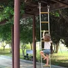 A young girl is climbing a yellow and white rope ladder hanging from a wooden structure in an outdoor park setting. Podgląd zdjęcia produktu