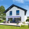 A modern white house with a blue and white striped awning over a patio door, a wooden deck with outdoor furniture, and a green lawn. Produktbild-Vorschau