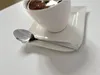 A white cup of coffee sits on a white saucer with a silver spoon resting on the saucer. Anteprima dell’immagine prodotto