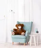 A large brown teddy bear sits in a light blue armchair in a room with white curtains and a floor lamp. Podgląd zdjęcia produktu