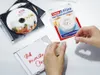 Hands using clear tape from a dispenser to attach a note to a CD case, with other CD cases and the tape packaging visible. Náhled obrázku produktu