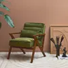 A green velvet armchair with a wooden frame is placed in a room with brown walls and a patterned rug. A plant and a framed artwork are visible in the background. Produktbild-Vorschau