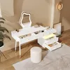 A white vanity table with a lit mirror and drawers, a stool, and a plant in a room. Voir les images du produit
