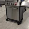 A close-up view of a grill with two cabinet doors and wheels. Náhled obrázku produktu