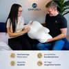 A man and a woman are sitting on a bed, holding and looking at an orthopedic pillow. The background has marketing text and icons. Produktbild-Vorschau
