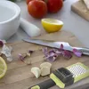 A kitchen scene with garlic, lemons, tomatoes, a mortar and pestle, a knife, and a garlic press on a wooden cutting board. Produktbild-Vorschau