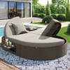 A circular rattan daybed with beige cushions is placed on a patio next to a swimming pool. The background shows a modern house with large glass doors and a manicured garden. Podgląd zdjęcia produktu