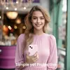 A woman is holding an iPhone in a pink case, smiling at the camera. The background is blurred, suggesting an outdoor or indoor setting with lights. Produktbild-Vorschau