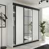 A modern white wardrobe with black accents and mirrored doors, set in a bright, contemporary room with a large window and plants. Produktbild-Vorschau