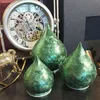 Three green glass teardrop-shaped decorative objects are displayed on a dark surface in front of a large clock. Produktbild-Vorschau