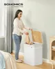 A woman is putting clothes into a white laundry basket with a bamboo lid. Produktbild-Vorschau