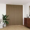 A taupe blackout curtain is shown hanging in a room, partially covering a window. A large potted plant is to the left of the curtain, and a wooden dresser is to the right. Náhľad obrázku produktu