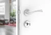 A close-up view of a silver door handle and lock mechanism on a white door, with a blurred interior room visible through the open doorway. Produktbild-Vorschau