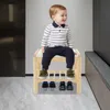 A young child sits on a wooden shoe bench with shoes on the lower shelves. Podgląd zdjęcia produktu