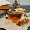 A glass mug filled with tea, garnished with orange slices, cinnamon sticks, and star anise, sits on a newspaper with cinnamon sticks tied with twine beside it. Books and a decorative jar are in the background. Voir les images du produit