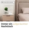 A white 3-in-1 wireless charging station is shown on a bedside table next to a plant, with a bed in the background. Produktbild-Vorschau