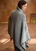 A woman wearing a gray knitted poncho stands in a room with wooden walls and floor. Produktbild-Vorschau