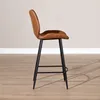 A side view of a brown leather bar stool with black metal legs against a light gray wall and a wooden floor. Produktbild-Vorschau