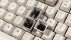 A close-up view of a keyboard with some keycaps removed, revealing small black figures inside the exposed switches. Produktbild-Vorschau