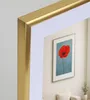 A framed picture of a red poppy on a blue background, displayed in a living room setting with a sofa and a plant. Produktbild-Vorschau