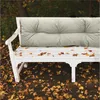 A white bench with a beige cushion sits outdoors surrounded by fallen autumn leaves. Náhled obrázku produktu