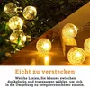 Close-up of string lights with bubble-like bulbs, some on greenery and some on a wooden surface. Náhled obrázku produktu